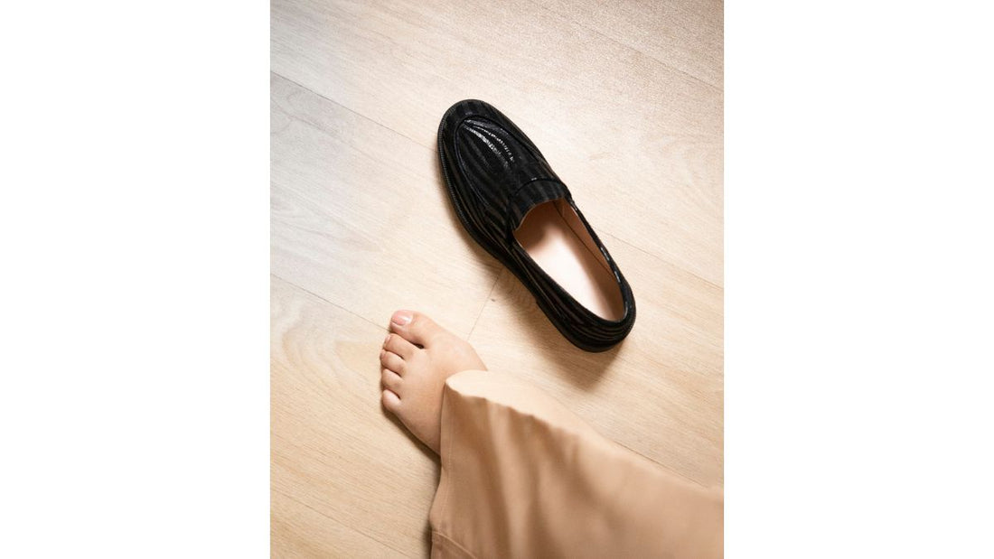 Mocassin noir en cuir Mouscin posé à côté d'un pied nu de femme sur un parquet en bois clair.