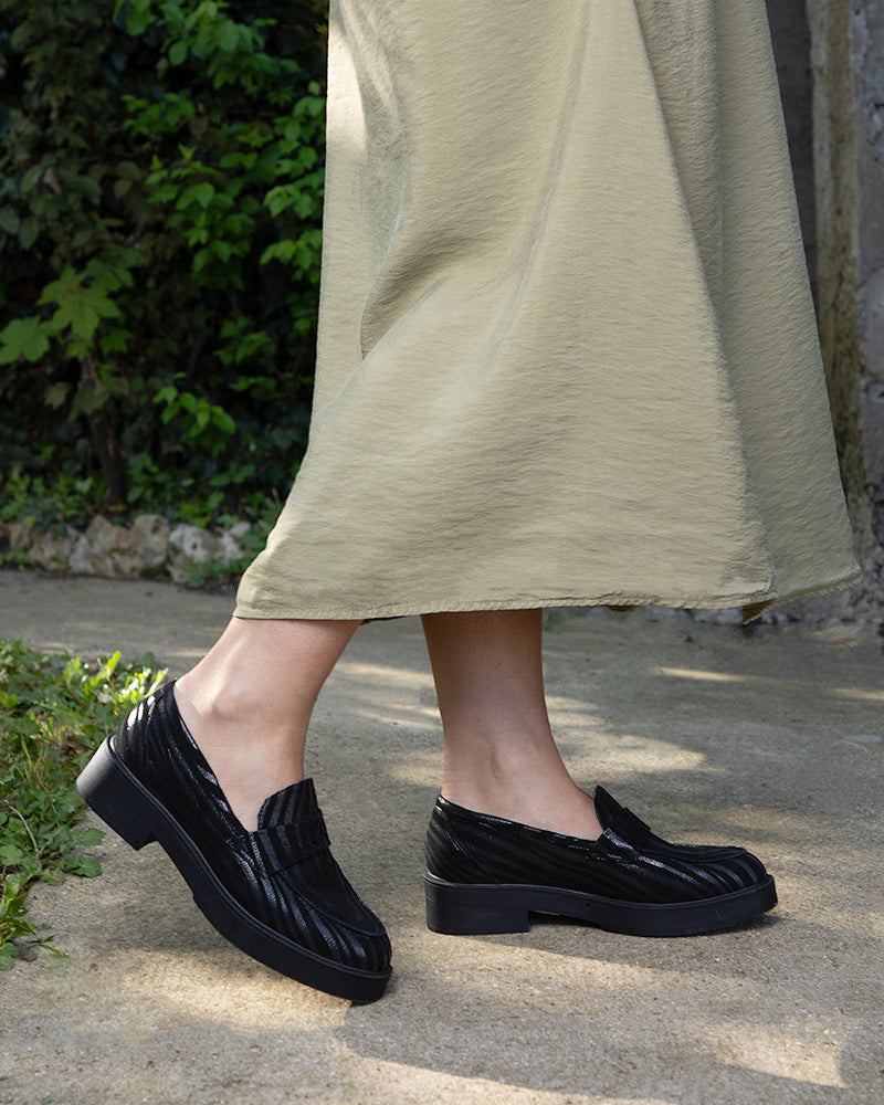 Gros plan sur les pieds d'une femme portant les mocassins à bout amande MOUSCIN : une marche confortable en extérieur sur du gravier qui illustre la souplesse du cuir noir texturé et l'espace généreux de la semelle plateforme, offrant un confort total sans aucune compression.